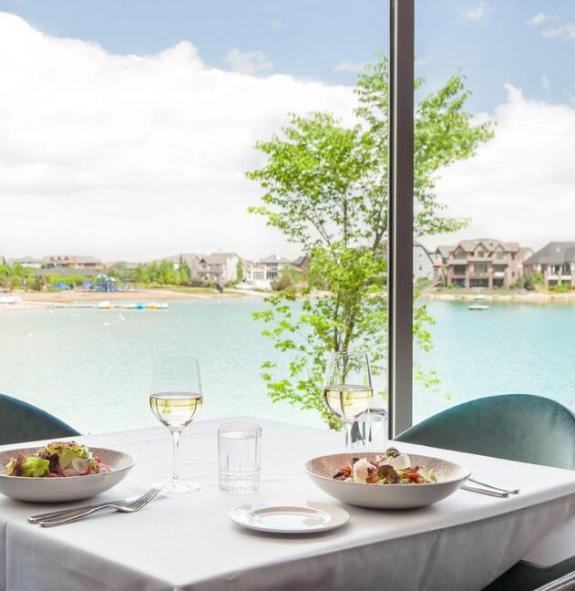 Lakeside dining view with gourmet meals and wine on a table, overlooking serene waters.