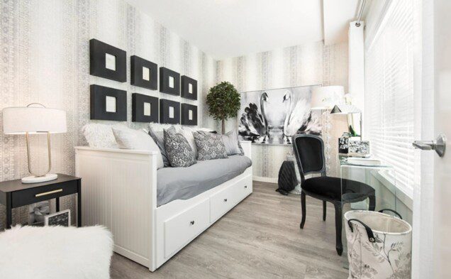 A contemporary 1-bedroom + den suite elegantly styled with a blend of modern aesthetics and timeless charm. The bedroom features a patterned accent wall, setting the backdrop for a symmetrical arrangement of black square frames. The daybed, in soothing gray tones, is adorned with patterned and textured pillows, emphasizing comfort and style. Storage drawers beneath provide practicality to the design.