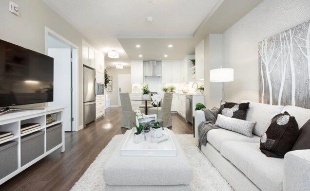 A spacious 2-bedroom suite featuring a living area with a mounted TV, a white sofa with decorative pillows, and a silver tree art piece on the wall. The open-concept kitchen boasts white cabinetry, stainless steel appliances, and a dining area with upholstered chairs around a flower centerpiece.