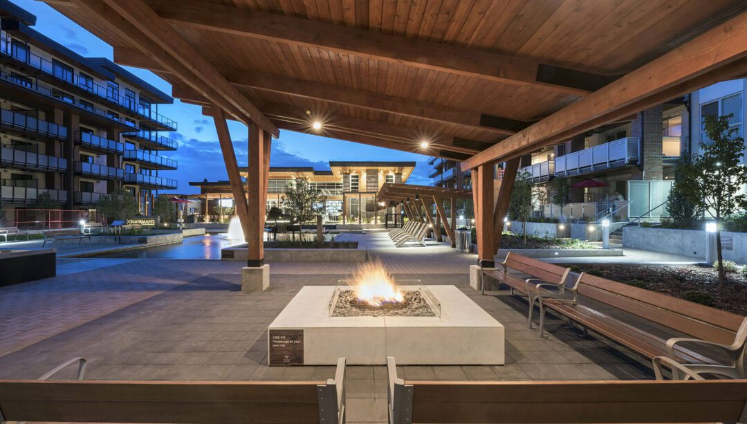 An inviting outdoor courtyard at twilight within the Journey Club complex, featuring a central fire pit with a robust flame. The space is surrounded by wooden benches and covered by a large wooden pergola structure, with ambient lighting adding to the warm atmosphere.