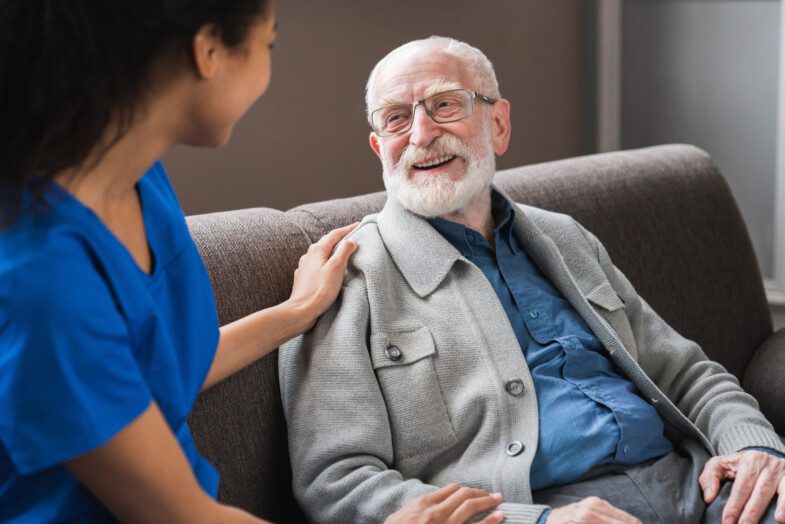 A nurse in blue scrubs comforting a smiling elderly man with glasses, seated on a couch.