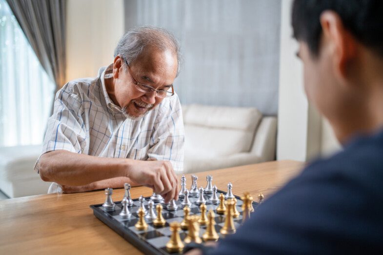 An elderly man with glasses smiling while playing chess with a younger opponent.