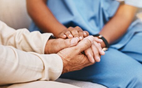 A caregiver in blue scrubs holds the hands of an elderly person, showing comfort and care.