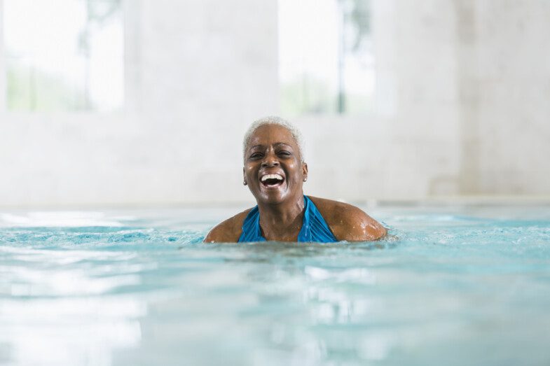 An elderly woman in a blue swimsuit, smiling and enjoying a swim in an indoor pool.